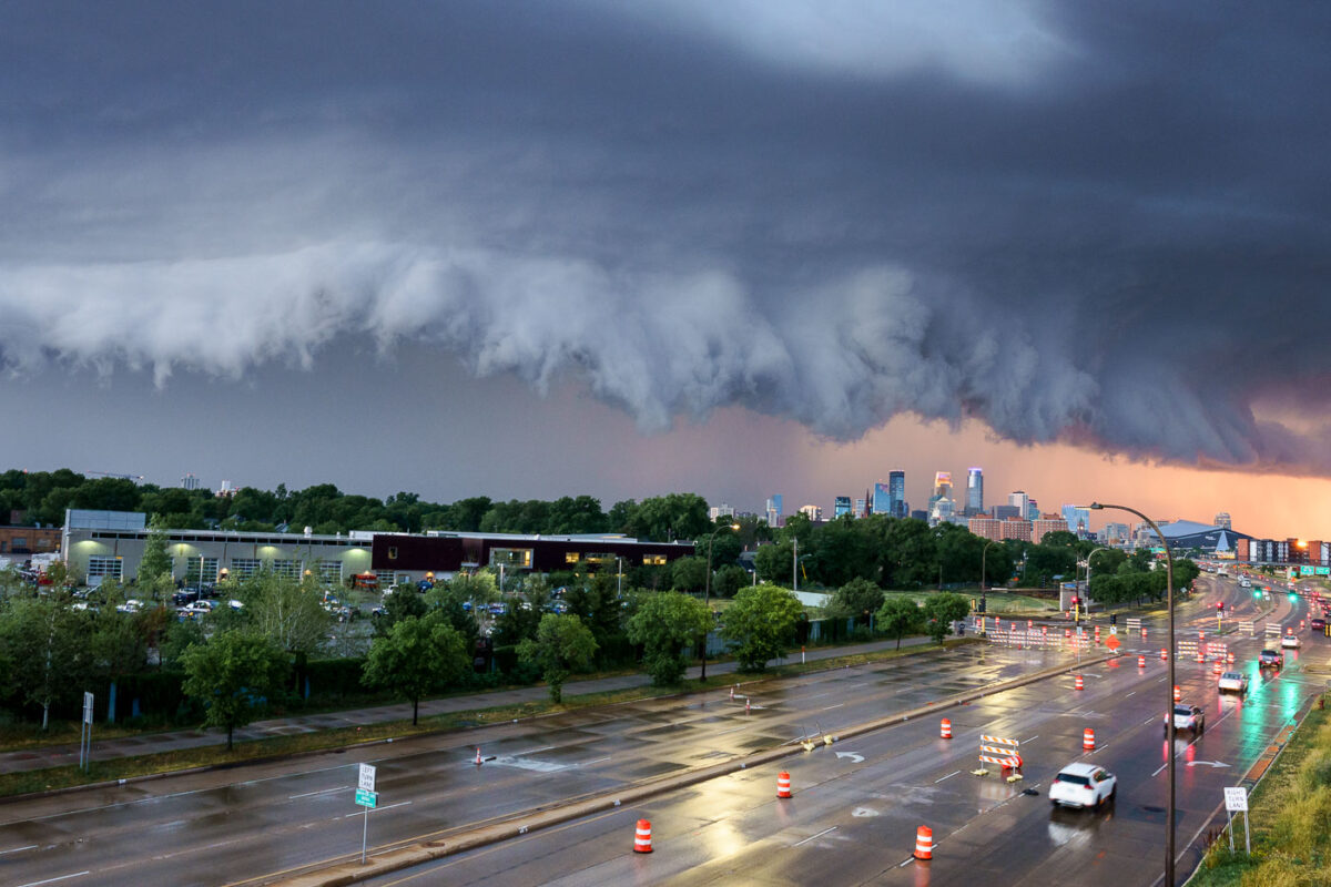A severe thunderstorm approaches the Minneapolis skyline on July 12, 2022. The city's downtown core, with its characteristic modern architecture, is visible in the distance under a dramatic, dark cloud formation. The foreground shows a wet roadway, indicating recent rainfall, with traffic continuing despite the approaching weather. This image captures a moment of atmospheric intensity over a major Midwestern urban center, highlighting the dynamic relationship between weather and the built environment.