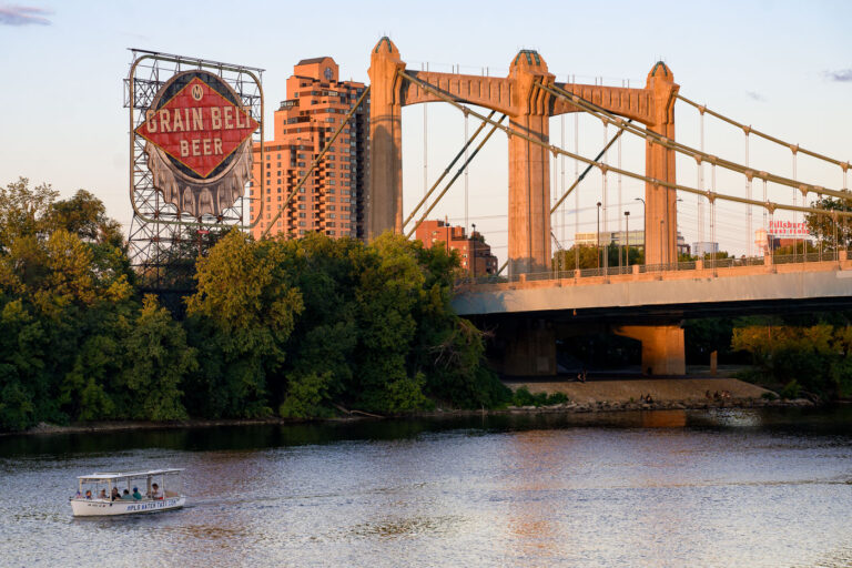 Minneapolis Water Taxi, Hennepin Ave Bridge, Grain Belt Sign 4 A water taxi travels on the Mississippi River in Minneapolis, passing under the Hennepin Avenue Bridge. Completed in 1929, the bridge is a significant example of early 20th-century infrastructure. The historic Grain Belt Beer sign, a remnant of the area's industrial past, is visible in the background, alongside modern residential buildings. The Mississippi River remains a vital transportation corridor, with services like the water taxi continuing its historical role.