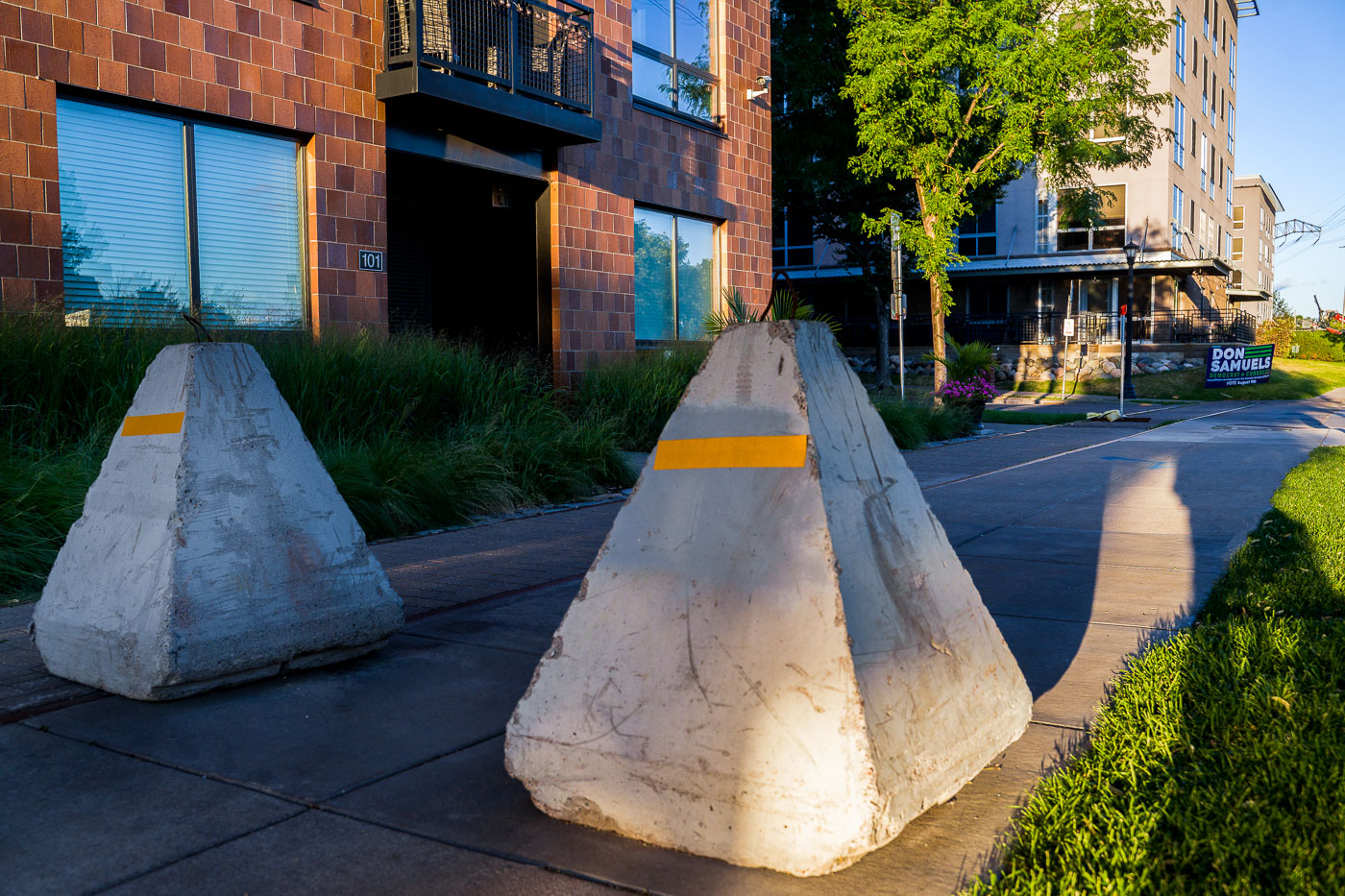 Concrete barricades are placed near the Stone Arch Bridge in Minneapolis, July 2022. These barriers were installed following public safety concerns and incidents that occurred on July 4th, 2022, a date often associated with large gatherings and past unrest in the city. The presence of these security measures reflects ongoing efforts to manage public spaces and ensure safety during periods of heightened activity. A campaign sign for Don Samuels, a candidate for Congress, is visible in the background, indicating the political climate of the area at the time.