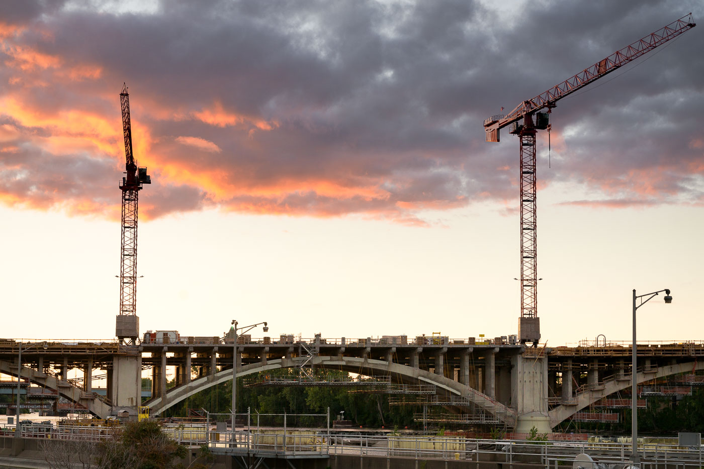 Construction cranes are visible over the Third Avenue Bridge in Minneapolis during a sunset. This bridge is a critical link between downtown Minneapolis and the Northeast community, spanning the Mississippi River. The ongoing reconstruction project aims to modernize the structure, which was originally built in the early 20th century and has served as a key piece of the city's transportation network.