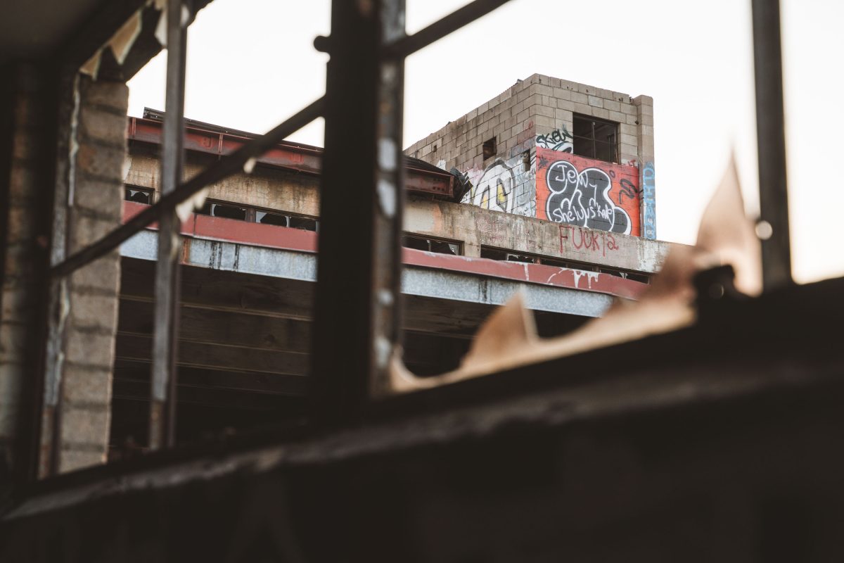Graffiti-covered brick building with red metal framework viewed through an industrial metal fence at abandoned Fruen Mill