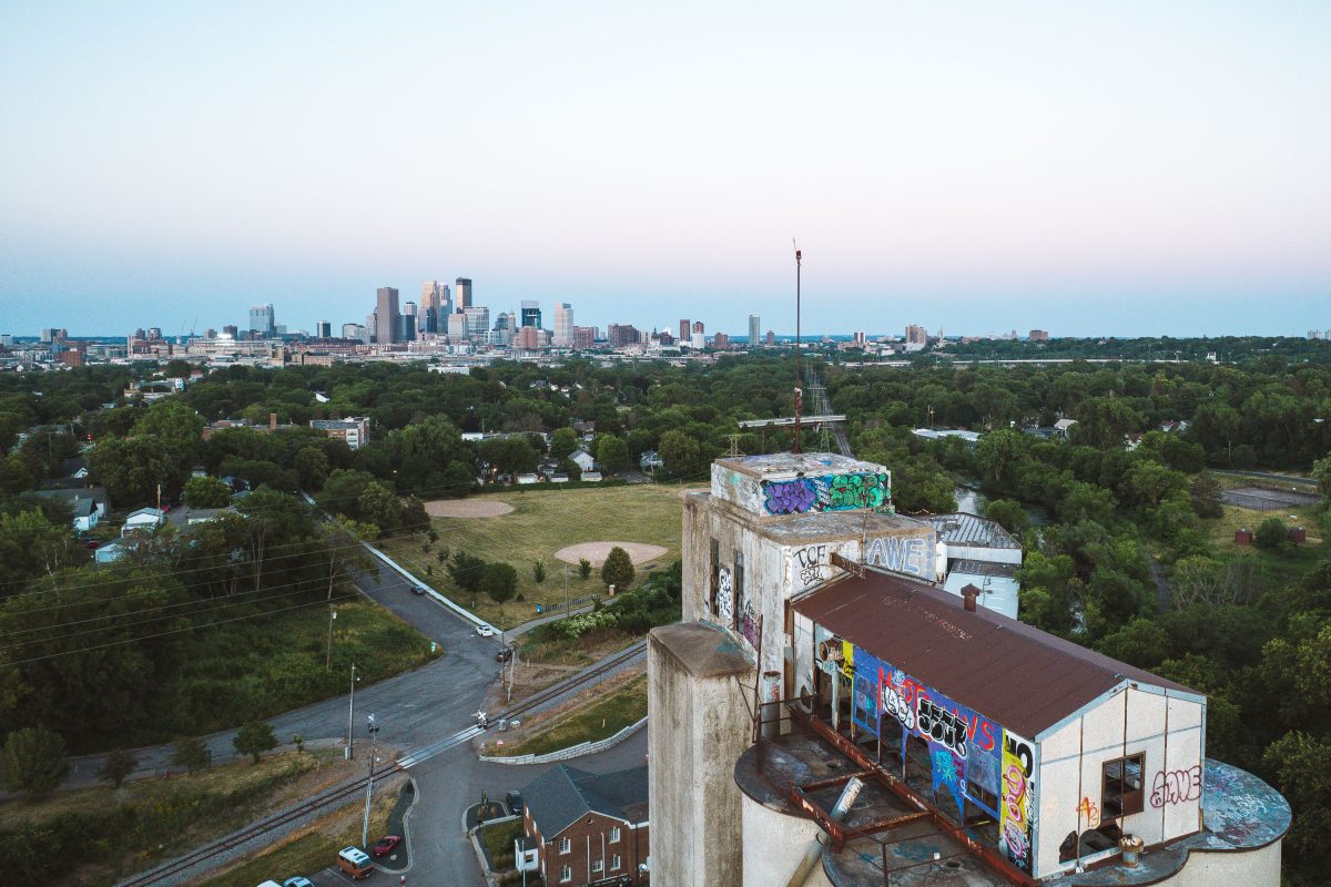 Graffiti-covered abandoned Fruen Mill complex with Minneapolis skyline visible across forested residential area.