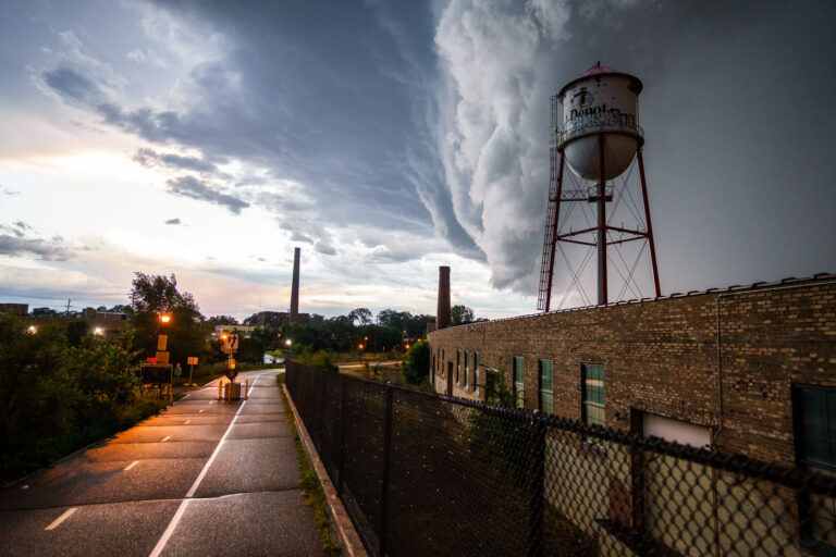 Minneapolis Depot Water Tower Under Storm Clouds 4 A massive storm front approaches Minneapolis on July 12, 2022, casting dark clouds over the city's industrial landscape. The photograph features a prominent water tower, labeled "Depot," and a brick industrial building, likely part of a former manufacturing or processing facility given the presence of smokestacks in the background. Water towers like this were essential for providing water pressure for industrial processes and fire suppression in urban and industrial areas. This particular tower, with its distinctive "Depot" marking, may have served a specific industrial complex or transportation hub within Minneapolis.