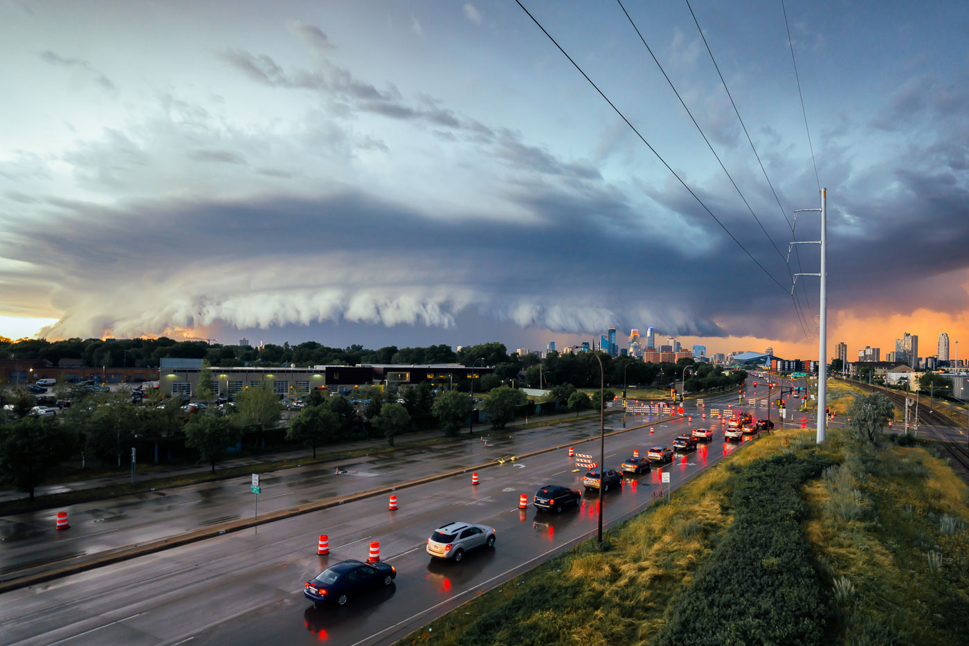 A powerful storm front approaches Minneapolis, Minnesota, on July 12, 2022. The image captures the city's skyline under a dramatic sky, with traffic moving along a wet highway. The presence of construction barrels suggests ongoing infrastructure work, a common characteristic of urban environments. This type of weather event is typical for the region during the summer months, impacting daily life and transportation.