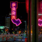 A bright magenta-and-red “PARK” marquee glows above 5th Street in downtown Minneapolis, its arrow pointing toward one of the city’s long-running parking ramps. The large mirrored surface on the right captures a full duplicate of the sign, emphasizing the heavy neon presence that once defined the downtown entertainment and retail district. Signs like this were installed in the mid-20th century as automobile traffic surged and developers converted older commercial buildings into multi-level garages to serve Nicollet Mall, the Gateway renewal area, and the growing high-rise core.

At street level, the rows of construction barrels and lane closures reflect Minneapolis’s ongoing cycle of roadway and transit upgrades — a pattern familiar in this part of the city as utility work, sidewalk rebuilds, and streetcar-era infrastructure get modern replacements. Even with the constant change, the surviving neon parking signs remain some of the most recognizable visual anchors of the nighttime downtown streetscape.