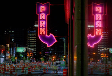 A bright magenta-and-red “PARK” marquee glows above 5th Street in downtown Minneapolis, its arrow pointing toward one of the city’s long-running parking ramps. The large mirrored surface on the right captures a full duplicate of the sign, emphasizing the heavy neon presence that once defined the downtown entertainment and retail district. Signs like this were installed in the mid-20th century as automobile traffic surged and developers converted older commercial buildings into multi-level garages to serve Nicollet Mall, the Gateway renewal area, and the growing high-rise core.

At street level, the rows of construction barrels and lane closures reflect Minneapolis’s ongoing cycle of roadway and transit upgrades — a pattern familiar in this part of the city as utility work, sidewalk rebuilds, and streetcar-era infrastructure get modern replacements. Even with the constant change, the surviving neon parking signs remain some of the most recognizable visual anchors of the nighttime downtown streetscape.