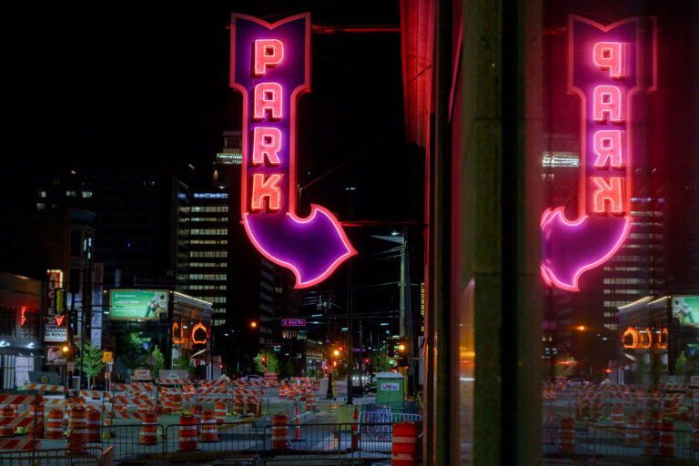Reflected Neon “PARK” Sign on 5th Street, Minneapolis 2 A bright magenta-and-red “PARK” marquee glows above 5th Street in downtown Minneapolis, its arrow pointing toward one of the city’s long-running parking ramps. The large mirrored surface on the right captures a full duplicate of the sign, emphasizing the heavy neon presence that once defined the downtown entertainment and retail district. Signs like this were installed in the mid-20th century as automobile traffic surged and developers converted older commercial buildings into multi-level garages to serve Nicollet Mall, the Gateway renewal area, and the growing high-rise core.At street level, the rows of construction barrels and lane closures reflect Minneapolis’s ongoing cycle of roadway and transit upgrades — a pattern familiar in this part of the city as utility work, sidewalk rebuilds, and streetcar-era infrastructure get modern replacements. Even with the constant change, the surviving neon parking signs remain some of the most recognizable visual anchors of the nighttime downtown streetscape.