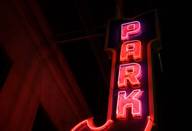 A classic vertical “PARK” marquee glows in red and magenta neon, marking one of downtown Minneapolis’s older parking structures that date back to the mid-20th century. These illuminated signs were once a staple of the city’s core, guiding drivers through the dense commercial grid before digital wayfinding and modern garages became standard. Many of these vintage markers survive as quiet architectural artifacts, attached to early high-rise buildings constructed during the postwar retail boom. Their hand-bent tubing and heavy steel housings reflect a period when neon craftsmanship was central to the city’s nighttime identity, especially along corridors near Nicollet Avenue, Hennepin Avenue, and the warehouse district.