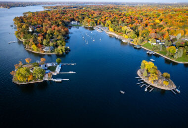 A high-elevation view over Lake Minnetonka captures several of the lake’s narrow peninsulas and sheltered bays at peak autumn color. The lake’s fragmented shoreline—shaped by glacial meltwater during the last Ice Age—creates dozens of protected inlets that became ideal sites for marinas, sailing clubs, and early resort development in the late 19th and early 20th centuries.

In this part of the lake, the docks and clustered sailboats reflect the area’s long-standing boating culture, while the densely wooded neighborhoods show how residential development was built around access to quiet coves rather than wide-open water. The surrounding canopy, shifting from green to deep red and gold, highlights why Lake Minnetonka remains one of the most photographed fall landscapes in the Twin Cities region.