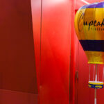 A Cupcake Vineyards promotional hot-air-balloon display stands at the edge of a red-lit entryway, its bright yellow and deep blue panels catching the saturated glow from the surrounding walls. These inflatable point-of-sale displays are common in liquor stores and bar entry corridors, designed to stand out under mixed lighting and draw attention to featured brands. The scene’s heavy red cast—likely from LED wash lighting or a painted interior corridor—creates a striking contrast with the cooler tones of the balloon, giving the space a graphic, almost stage-set quality often found in Minneapolis nightlife districts where narrow passages and dramatic lighting create strong visual color blocks.