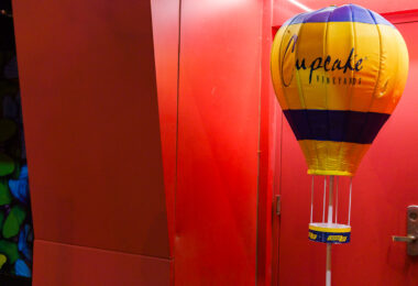 A Cupcake Vineyards promotional hot-air-balloon display stands at the edge of a red-lit entryway, its bright yellow and deep blue panels catching the saturated glow from the surrounding walls. These inflatable point-of-sale displays are common in liquor stores and bar entry corridors, designed to stand out under mixed lighting and draw attention to featured brands. The scene’s heavy red cast—likely from LED wash lighting or a painted interior corridor—creates a striking contrast with the cooler tones of the balloon, giving the space a graphic, almost stage-set quality often found in Minneapolis nightlife districts where narrow passages and dramatic lighting create strong visual color blocks.