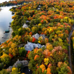 This aerial view follows the wooded shoreline of Deephaven, a small lakeside community on the southeast edge of Lake Minnetonka. The neighborhood developed around a combination of 19th-century summer cottages and later year-round homes, many of which were built on large forested lots that still preserve much of the area’s original tree cover. In autumn, the mix of maple, oak, and basswood trees creates a dense canopy of reds, yellows, and orange tones that nearly conceals the homes and winding shoreline roads below. Deephaven’s bay-front docks, protected inlets, and narrow peninsulas are distinctive features of this part of Minnetonka, reflecting the lake’s long history as a regional retreat and residential enclave.
