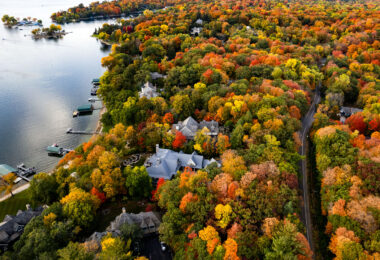 This aerial view follows the wooded shoreline of Deephaven, a small lakeside community on the southeast edge of Lake Minnetonka. The neighborhood developed around a combination of 19th-century summer cottages and later year-round homes, many of which were built on large forested lots that still preserve much of the area’s original tree cover. In autumn, the mix of maple, oak, and basswood trees creates a dense canopy of reds, yellows, and orange tones that nearly conceals the homes and winding shoreline roads below. Deephaven’s bay-front docks, protected inlets, and narrow peninsulas are distinctive features of this part of Minnetonka, reflecting the lake’s long history as a regional retreat and residential enclave.