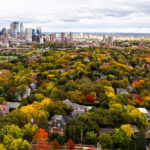 A wide aerial view looks east toward the Minneapolis skyline, rising above the dense canopy of the Kenwood and Lowry Hill neighborhoods during peak autumn color. This part of the city was developed in the late 19th and early 20th centuries as a streetcar suburb, designed around curving streets, large residential lots, and proximity to the Chain of Lakes. Many of the homes below are early Minneapolis mansions built by lumber and milling-era industrialists, now tucked beneath mature trees that give the neighborhood one of the city’s most dramatic fall transitions.

In the middle distance, the Basilica of Saint Mary, Loring Park, and the early downtown warehouse district reflect Minneapolis’s layered growth—from industrial rail corridors to modern glass towers. The contrast between the dense urban core and the tree-covered residential districts highlights how closely the city’s historic neighborhoods were shaped by parks, lakes, and the parkway system planned by Theodore Wirth in the early 1900s.