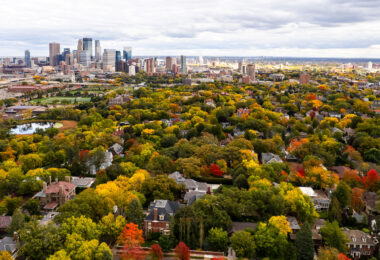 A wide aerial view looks east toward the Minneapolis skyline, rising above the dense canopy of the Kenwood and Lowry Hill neighborhoods during peak autumn color. This part of the city was developed in the late 19th and early 20th centuries as a streetcar suburb, designed around curving streets, large residential lots, and proximity to the Chain of Lakes. Many of the homes below are early Minneapolis mansions built by lumber and milling-era industrialists, now tucked beneath mature trees that give the neighborhood one of the city’s most dramatic fall transitions.

In the middle distance, the Basilica of Saint Mary, Loring Park, and the early downtown warehouse district reflect Minneapolis’s layered growth—from industrial rail corridors to modern glass towers. The contrast between the dense urban core and the tree-covered residential districts highlights how closely the city’s historic neighborhoods were shaped by parks, lakes, and the parkway system planned by Theodore Wirth in the early 1900s.