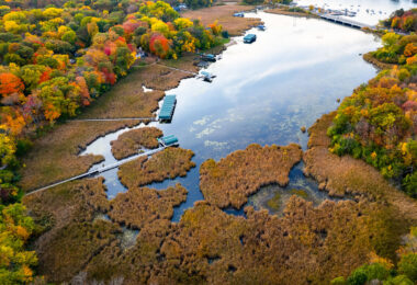 This aerial view looks over one of Lake Minnetonka’s shallow eastern bays, where a broad wetland of cattails and floating vegetation meets a line of private docks and covered boat slips. These marshy inlets are among the lake’s most ecologically important areas, filtering runoff, supporting migratory birds, and preserving habitat that once covered far more of the shoreline before 20th-century dredging and residential development. The fall canopy surrounding the bay shows the mix of hardwoods common in the Deephaven and Greenwood areas, with red maples, oaks, and ash trees creating a dense belt of color around the water. Farther in the distance, the channel and bridge leading toward Carson’s Bay hint at the lake’s extensive network of connected basins, many of which were linked by canals built in the late 1800s to support early resort traffic.