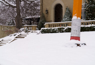 A large pencil sculpture stands partly buried in fresh snow near Lake of the Isles in Minneapolis. The piece—well-known to neighborhood walkers and cyclists—sits on the lawn of a private residence and has become a quiet local landmark. Framed by stone steps, evergreens, and early-season snowfall, it reflects the eclectic public-facing art found throughout the surrounding residential areas of the Chain of Lakes.