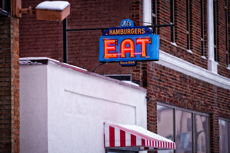 Al’s Hamburgers Neon Sign in Winter, Green Bay 4 The glowing neon “EAT” sign at Al’s Hamburgers stands out against the snowy backdrop of winter in Minneapolis. The sign, with its bright mid-century colors and bold lettering, has been part of the business’s identity since the diner opened in 1934. Its classic styling reflects the era when small neighborhood restaurants relied on eye-catching storefront signs to draw in passing traffic. Light snow gathers on the striped awning below, adding a seasonal layer to one of the area’s longstanding local landmarks.