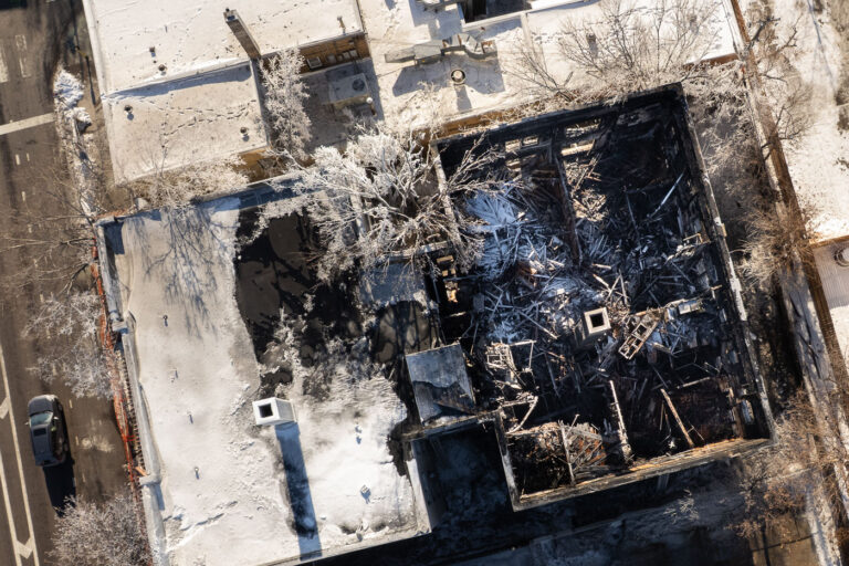Lyndale Avenue Fire Ruins from Above 1 An aerial view shows the charred remains of a vacant apartment building on Lyndale Avenue in Minneapolis the morning after a fire swept through the structure. Snow outlines the building’s footprint and surrounding rooftops, contrasting sharply with the blackened debris and collapsed interior walls. Frozen trees, scorched timbers, and scattered structural fragments highlight both the intensity of the blaze and the rapid onset of winter conditions that followed.