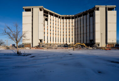A partially dismantled mid-century office tower stands open to the winter light in Bloomington, Minnesota, as demolition crews continue taking down the long-vacant structure. The building’s exposed interior and collapsed debris field reflect the broader wave of suburban redevelopment that has reshaped commercial corridors near I-494 and the Minneapolis–St. Paul International Airport. Heavy equipment rests at the base of the tower, marking another step in clearing the site for future use amid changing regional land-use patterns.