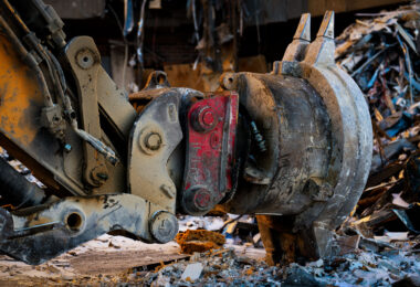 A close-up view of a hydraulic excavator’s quick-coupler and demolition attachment during the teardown of the former Southgate Office Tower in Bloomington, Minnesota. The worn steel, hydraulic lines, and layers of dust and debris show the heavy strain placed on equipment during large-scale structural demolition. Piles of shattered concrete, insulation, and twisted metal surrounding the attachment reflect the ongoing dismantling of the mid-century office complex.