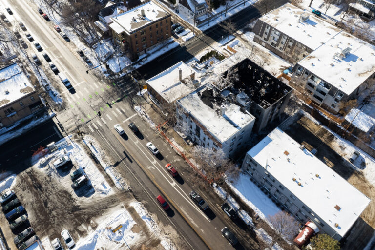 Lyndale Avenue Fire Aftermath from Above 4 An aerial view of the fire-damaged apartment building along Lyndale Avenue in Minneapolis, photographed the morning after the incident. The structure’s roof is completely gone, and its upper floors are visibly burned out, leaving only the exterior walls and charred framing behind. Surrounding buildings—mostly mid-century apartment blocks—show normal winter conditions, their roofs covered in fresh snow. Traffic moves through the busy Lyndale and 24th Street intersection while the fire scene remains cordoned off with temporary fencing. The image highlights the dense residential fabric of this part of the city and the stark contrast between the intact neighborhood and the gutted building at the center of the block.