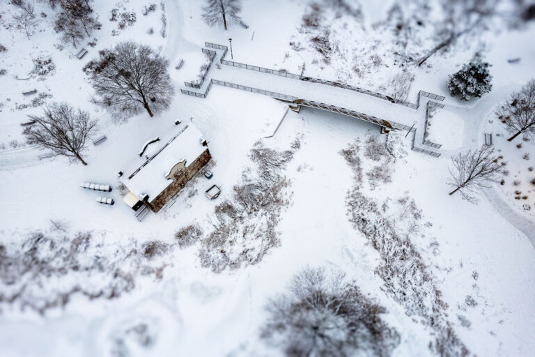 Loring Park Footbridge After Fresh Snowfall 3 A fresh snowfall settles over Loring Park near downtown Minneapolis, softening the landscape around the park’s pedestrian bridge and historic shelter building. From above, the snow highlights the geometric layout of paths, railings, and terraces surrounding the bridge, which crosses a narrow channel connecting sections of the park’s pond system. Loring Park—a key part of the city’s late-19th-century park plan—remains one of Minneapolis’s most recognizable urban green spaces, bordered by the Loring Greenway and the southern edge of downtown. The new snow creates a uniform winter texture across the lawns, benches, and shoreline vegetation, with only light footprints and plowed walkways hinting at early morning activity in the park.