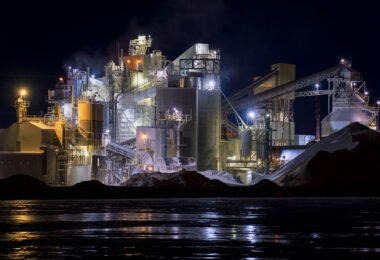 The Graymont lime processing plant along the Fox River in Green Bay, Wisconsin, illuminated during nighttime operations. The facility’s network of silos, conveyors, and dust-collection towers stands out against the dark sky, with steam drifting from active kilns and processing units. Reflections from the plant’s industrial lighting shimmer across the frozen river surface, highlighting the site’s role in the region’s long-standing manufacturing and mineral production corridor.