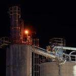 Lime Silos and Transfer Systems at Night 3 Illuminated by a single amber work light, the upper platforms and transfer pipes of the Graymont lime plant in Green Bay, Wisconsin stand out against the night sky. The cylindrical storage silos are connected by enclosed conveyor and piping systems that move processed lime between different stages of production. Catwalks, ladders, and pipe supports trace the geometry of an industrial site that has long supplied lime for regional steelmaking, water treatment, and construction uses.