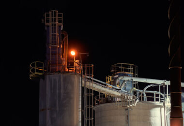 Illuminated by a single amber work light, the upper platforms and transfer pipes of the Graymont lime plant in Green Bay, Wisconsin stand out against the night sky. The cylindrical storage silos are connected by enclosed conveyor and piping systems that move processed lime between different stages of production. Catwalks, ladders, and pipe supports trace the geometry of an industrial site that has long supplied lime for regional steelmaking, water treatment, and construction uses.