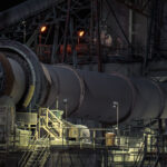 Rotary Kiln System at the Graymont Lime Plant 1 A section of the rotary kiln system at the Graymont lime plant in Green Bay, Wisconsin, captured at night under maintenance and work lights. The massive rotating kiln—central to the calcination process that transforms limestone into high-purity lime—runs along a network of elevated platforms, access stairs, and steel supports. The orange glow from overhead lamps highlights the heat-intensive nature of kiln operations, while the surrounding catwalks and conveyors reflect the layered engineering required to keep the continuous production line moving.