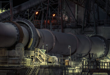 A section of the rotary kiln system at the Graymont lime plant in Green Bay, Wisconsin, captured at night under maintenance and work lights. The massive rotating kiln—central to the calcination process that transforms limestone into high-purity lime—runs along a network of elevated platforms, access stairs, and steel supports. The orange glow from overhead lamps highlights the heat-intensive nature of kiln operations, while the surrounding catwalks and conveyors reflect the layered engineering required to keep the continuous production line moving.