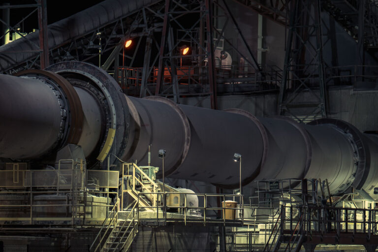 Rotary Kiln System at the Graymont Lime Plant 4 A section of the rotary kiln system at the Graymont lime plant in Green Bay, Wisconsin, captured at night under maintenance and work lights. The massive rotating kiln—central to the calcination process that transforms limestone into high-purity lime—runs along a network of elevated platforms, access stairs, and steel supports. The orange glow from overhead lamps highlights the heat-intensive nature of kiln operations, while the surrounding catwalks and conveyors reflect the layered engineering required to keep the continuous production line moving.
