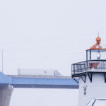 A CTS tractor-trailer crosses the Leo Frigo Memorial Bridge—locally known as the Tower Drive Bridge—during a winter snowstorm in Green Bay, Wisconsin. In the foreground, the familiar shingled lighthouse structure along the riverfront stands out against the white sky, its lantern room partially frosted as lake-effect snow drifts through the scene. The contrast between the elevated highway and the shoreline landmark highlights the mix of transportation corridors that define Green Bay’s industrial waterfront.
