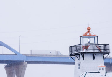 A CTS tractor-trailer crosses the Leo Frigo Memorial Bridge—locally known as the Tower Drive Bridge—during a winter snowstorm in Green Bay, Wisconsin. In the foreground, the familiar shingled lighthouse structure along the riverfront stands out against the white sky, its lantern room partially frosted as lake-effect snow drifts through the scene. The contrast between the elevated highway and the shoreline landmark highlights the mix of transportation corridors that define Green Bay’s industrial waterfront.