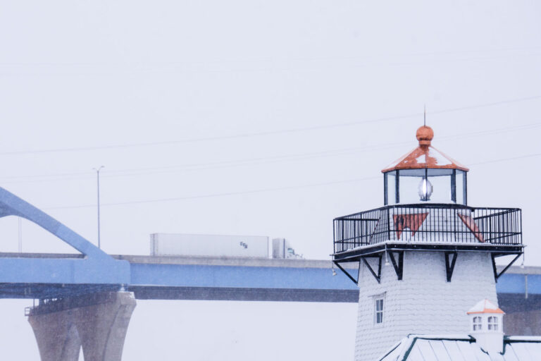 Winter Traffic on the Leo Frigo Bridge 3 A CTS tractor-trailer crosses the Leo Frigo Memorial Bridge—locally known as the Tower Drive Bridge—during a winter snowstorm in Green Bay, Wisconsin. In the foreground, the familiar shingled lighthouse structure along the riverfront stands out against the white sky, its lantern room partially frosted as lake-effect snow drifts through the scene. The contrast between the elevated highway and the shoreline landmark highlights the mix of transportation corridors that define Green Bay’s industrial waterfront.