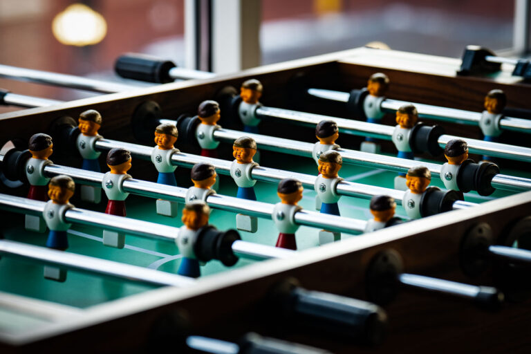 Foosball Table Figures in Afternoon Light 1 A close-up view of a classic foosball table shows rows of molded player figures lined up on chrome-plated rods, illuminated by soft afternoon light from a nearby window. The design—bright jerseys, simple facial features, and sturdy construction—reflects the durable style common in commercial and recreational tables since the mid-20th century. Often found in community centers, break rooms, and pubs, foosball tables like this one are built for fast play and social interaction, with standardized layouts that mirror the positioning of real soccer formations.