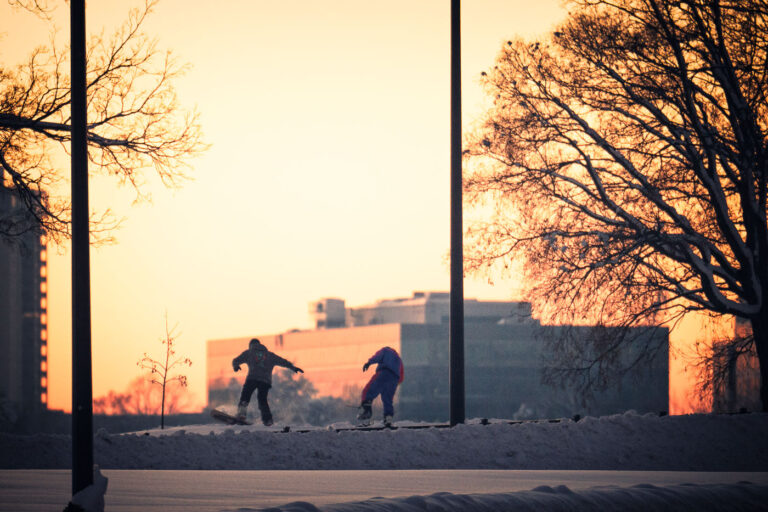 Sunset Snowboarding at Bde Maka Ska 2 Two snowboarders make use of fresh snow along the parkway near Bde Maka Ska in Minneapolis after a major winter storm. The lake—renamed in 2018 to restore its Dakota place name—anchors the Chain of Lakes system and serves as one of the city’s most heavily used recreation areas year-round. Heavy snowfall often transforms the surrounding trails and embankments into informal terrain for skiing, snowboarding, and winter biking, reflecting how quickly the area adapts to seasonal conditions. The early-morning light and cleared parkway signal the coordinated snow-response efforts that keep the city’s lakes district accessible even during severe weather.