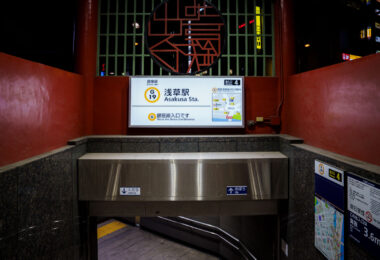 An entrance to Tokyo Metro’s Asakusa Station on the Ginza Line, marked with the station code G-19, leads commuters down a clean, tiled stairway under the red-painted gateway structure. The illuminated signage displays route maps and directional guidance, combining modern wayfinding with the surrounding area’s traditional aesthetic. Located near Sensō-ji Temple, this entrance reflects Asakusa’s balance of old Tokyo charm and contemporary transit design.