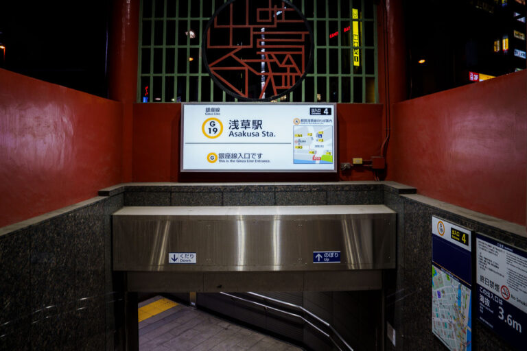 Asakusa Station Entrance Tokyo 4 An entrance to Tokyo Metro’s Asakusa Station on the Ginza Line, marked with the station code G-19, leads commuters down a clean, tiled stairway under the red-painted gateway structure. The illuminated signage displays route maps and directional guidance, combining modern wayfinding with the surrounding area’s traditional aesthetic. Located near Sensō-ji Temple, this entrance reflects Asakusa’s balance of old Tokyo charm and contemporary transit design.