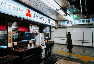 A small soba counter operates inside Asakusa Underground Street, one of Tokyo’s oldest surviving subterranean shopping arcades. Tucked beneath the streets near Asakusa Station, these compact eateries serve quick bowls of soba and udon to commuters and visitors moving between the metro and the surrounding neighborhood.

Opened in 1955, Asakusa Underground Street is considered the oldest underground shopping street in Japan. Its narrow corridors, low ceilings, and tightly packed storefronts preserve a postwar atmosphere that has largely vanished from Tokyo as major stations modernized. The passageway originally developed as a practical connection point for transit riders, but it evolved into a lively network of bars, snack counters, tobacconists, and small service shops. Today, despite gradual renovation, it remains one of the city’s most atmospheric relics of mid-20th-century urban life—an example of how Tokyo’s underground spaces doubled as both transportation infrastructure and neighborhood social hubs.