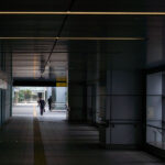 Business man in Tokyo Japan 3 A quiet, modern pedestrian corridor connects parts of a Japanese transit hub, its metallic walls and ceiling panels reflecting soft daylight from the far exit. Yellow tactile paving guides commuters toward the outside, where a brief view of greenery contrasts with the tunnel’s rigid geometry. The minimalist design and clean lines reflect Japan’s approach to functional urban infrastructure—precise, orderly, and human-scaled.