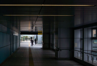A quiet, modern pedestrian corridor connects parts of a Japanese transit hub, its metallic walls and ceiling panels reflecting soft daylight from the far exit. Yellow tactile paving guides commuters toward the outside, where a brief view of greenery contrasts with the tunnel’s rigid geometry. The minimalist design and clean lines reflect Japan’s approach to functional urban infrastructure—precise, orderly, and human-scaled.
