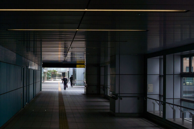Business man in Tokyo Japan 1 A quiet, modern pedestrian corridor connects parts of a Japanese transit hub, its metallic walls and ceiling panels reflecting soft daylight from the far exit. Yellow tactile paving guides commuters toward the outside, where a brief view of greenery contrasts with the tunnel’s rigid geometry. The minimalist design and clean lines reflect Japan’s approach to functional urban infrastructure—precise, orderly, and human-scaled.