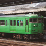 A classic green Japanese commuter train sits at a station platform, its utilitarian design and weathered paint evoking an earlier era of rail travel. These older local-line cars, once common throughout regional Japan, continue to serve smaller routes with reliability and simplicity. The muted tones of the train and station canopy, along with a uniformed conductor preparing for departure, create a nostalgic glimpse into the everyday rhythm of Japan’s vast and enduring railway network.