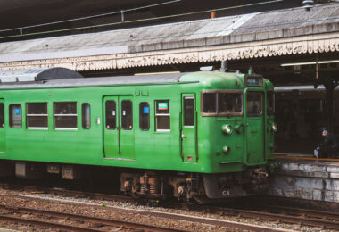 A classic green Japanese commuter train sits at a station platform, its utilitarian design and weathered paint evoking an earlier era of rail travel. These older local-line cars, once common throughout regional Japan, continue to serve smaller routes with reliability and simplicity. The muted tones of the train and station canopy, along with a uniformed conductor preparing for departure, create a nostalgic glimpse into the everyday rhythm of Japan’s vast and enduring railway network.