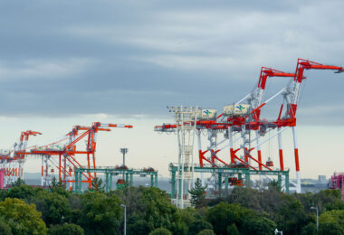 Towering gantry cranes line the waterfront at a Japanese container terminal, their red and white frames contrasting sharply against the overcast sky. The cranes, marked with “Evergreen” and “K” Line logos, are used to load and unload massive cargo ships that connect Japan to global trade routes. Surrounded by greenery and infrastructure, the scene reflects the blend of industry and environment common to Japan’s modern port cities—where precision logistics and advanced automation keep goods moving efficiently through one of the world’s busiest maritime networks.