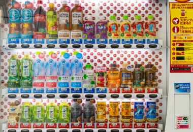 A brightly lit Japanese vending machine displays a meticulously arranged lineup of drinks—from Coca-Cola and Fanta to bottled teas, mineral waters, and Georgia coffee blends. Each row is clearly priced, some marked with blue “つめたい” (cold) or red “あったかい” (hot) labels, offering both chilled and warmed beverages. The right panel promotes the “Coke ON” smartphone app, merging tradition with tech-driven convenience. This snapshot captures Japan’s signature vending culture: accessible, efficient, and surprisingly aesthetic in its everyday precision.