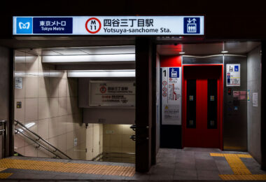 The entrance to Yotsuya-sanchome Station on Tokyo Metro’s Marunouchi Line (M11) is shown at night, featuring a clean, modern design with bright signage and red elevator doors. The station serves the Shinjuku district and connects to key destinations like Shinjuku, Ogikubo, and Tokyo Station. Braille paving and clear bilingual signs reflect Tokyo Metro’s accessibility standards and emphasis on wayfinding.
