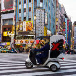 A delivery scooter moves through the large scramble crossing on Central Road in Kabukichō, one of Shinjuku’s busiest commercial corridors. This stretch of the district, located just north of Yasukuni-dōri, has been a dense entertainment zone since the postwar reconstruction era, eventually becoming known for its mix of cinemas, restaurants, arcades, karaoke towers, and neon advertising stacked across narrow mid-rise buildings.

The tall signs and narrow storefronts reflect the vertical commercial pattern typical of Shinjuku’s nightlife blocks, where multiple businesses occupy each floor and compete for visibility at street level. In the distance, the Godzilla head mounted atop the Hotel Gracery Shinjuku—installed in 2015 as part of the Toho Cinemas complex—has become one of the district’s most recognizable landmarks, connecting the area’s contemporary tourism appeal with Japan’s long-running film history.